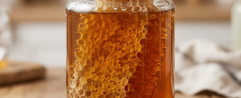 Glass jar of honey containing honeycomb on wooden table with honey dipper