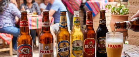 Six bottles of Bolivian beer varieties lined up on a wooden table with snacks and a glass being filled