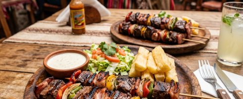 Grilled meat and vegetable skewers with salad, fries, dipping sauce, and a drink on wooden plates