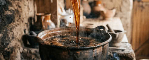 A hand holding a wooden ladle pouring a dark liquid from a boiling pot on an open fire