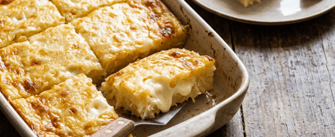 Golden cheesy cassava bake in a casserole dish with a slice served on a plate alongside lime wedges and slaw