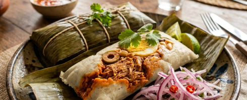 Savory tamale wrapped in banana leaf accompanied by pickled red onions, lime, and salsa